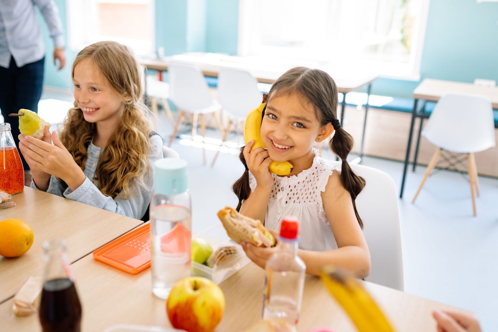 Two smiling girls enjoying snacks during break time in a school classroom.