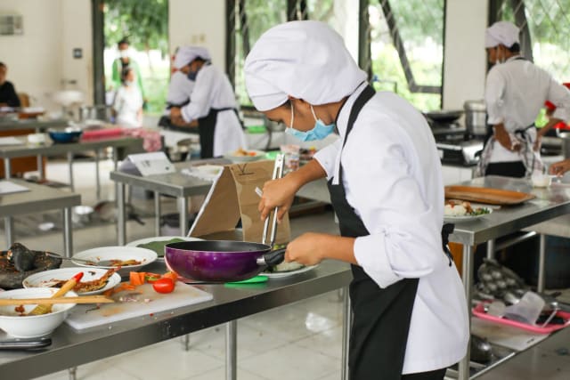 Culinary students in uniforms practice cooking skills in a professional kitchen setting.